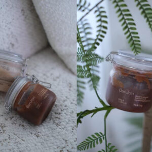 Brown body scrub jars displayed on stone with green foliage.
