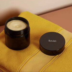 Open dark BunJoin body butter jar on a mustard-colored towel.