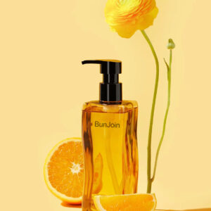 Golden BunJoin skin oil pump bottle beside orange slices and a yellow flower.