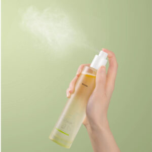 Hand spraying a clear body mist bottle against a pale green background.